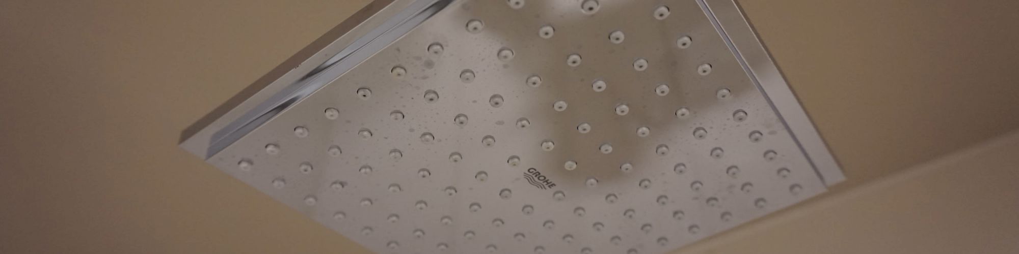 The image shows a square-shaped chrome shower head installed on a bathroom ceiling. The shower head has many small water holes.