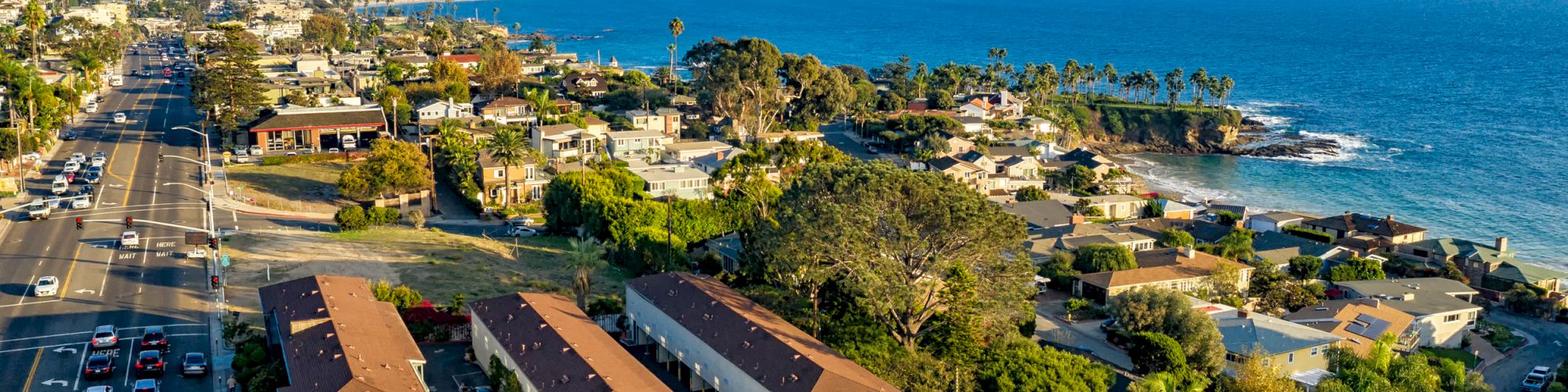 A coastal neighborhood with houses alongside a busy road, next to the ocean under a blue sky.