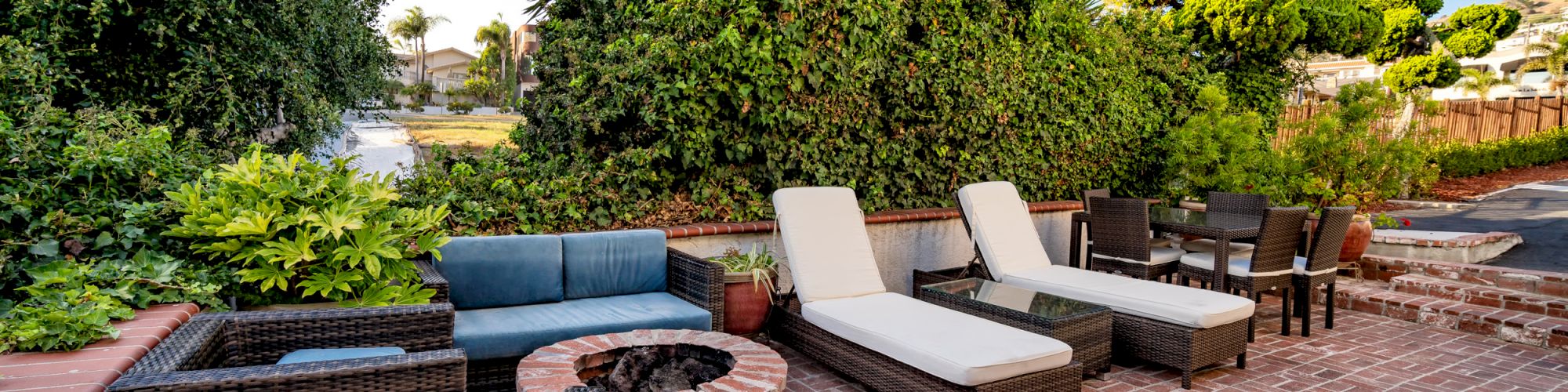 An outdoor patio with wicker furniture, loungers, a table, and chairs on a brick surface surrounded by lush greenery and clear skies.