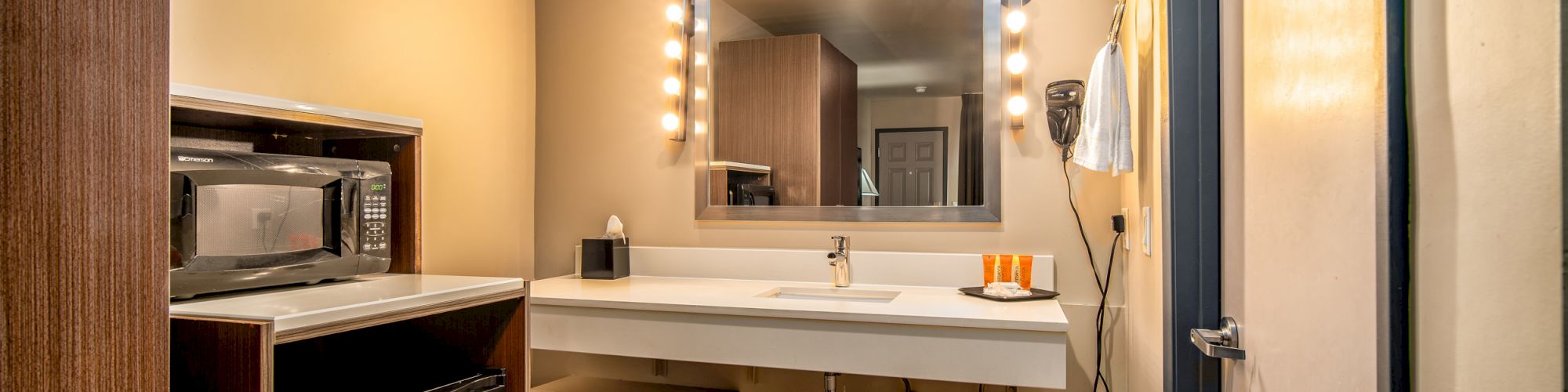 This image shows a bathroom with a lit mirror, a sink, a wall-mounted hairdryer, a microwave, and a mini-fridge, all under warm lighting.