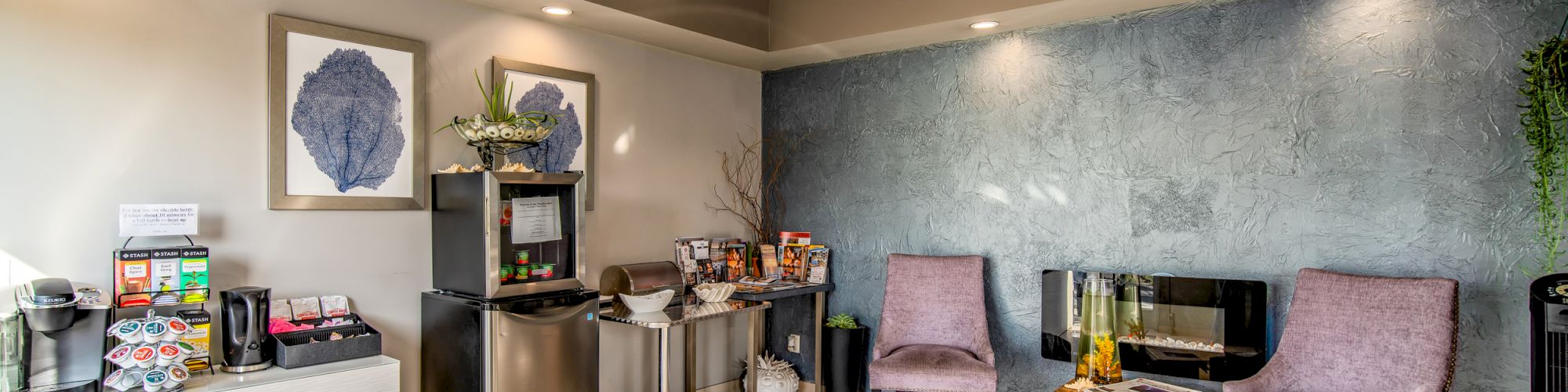 A cozy seating area with two chairs, a small table, coffee station, mini-fridge, snacks, and decorative wall art. Natural light illuminates the space.