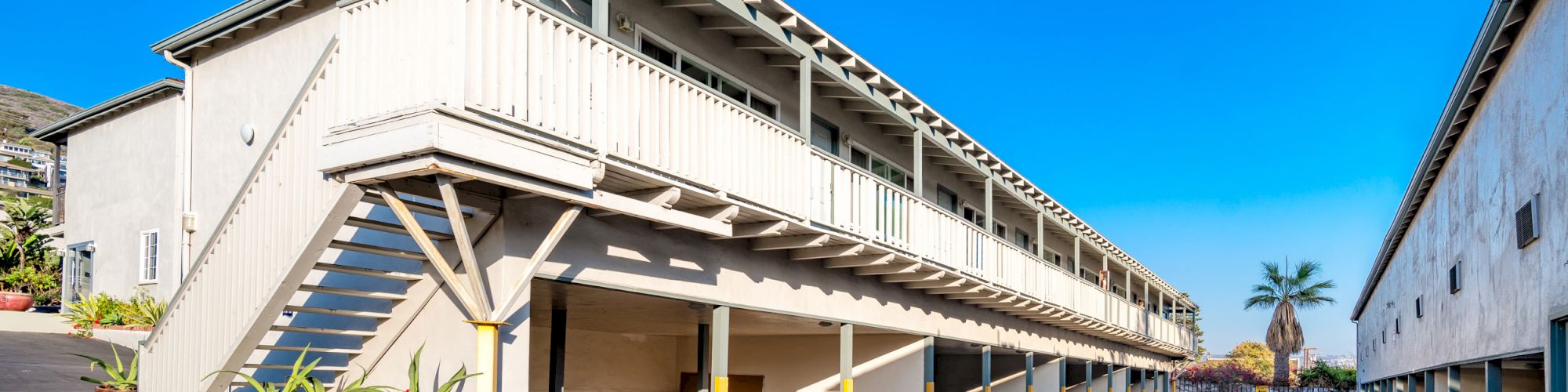 The image shows an exterior view of a two-story apartment building with a staircase, overhang parking, and a clear blue sky.