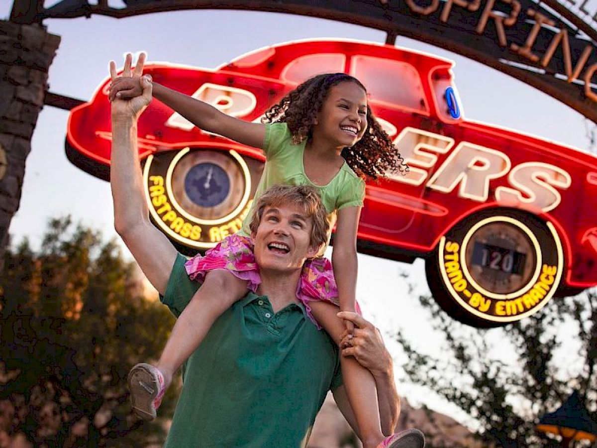 A happy man carrying a joyful girl on his shoulders in front of a sign featuring a red car and amusement park theming.