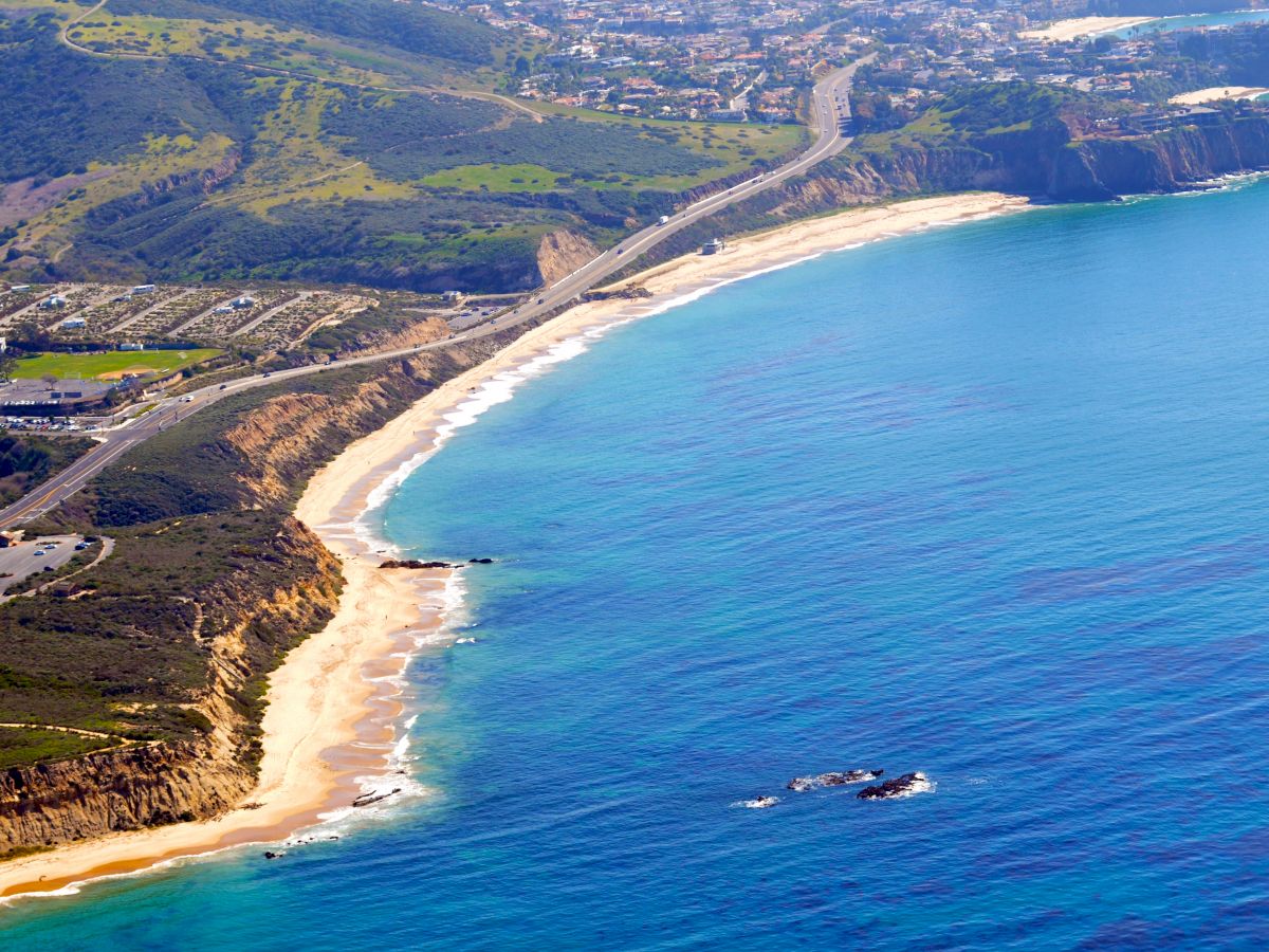 An aerial view of a coastal area with a winding road, sandy beaches, green hills, and clear blue waters stretching along the shoreline.