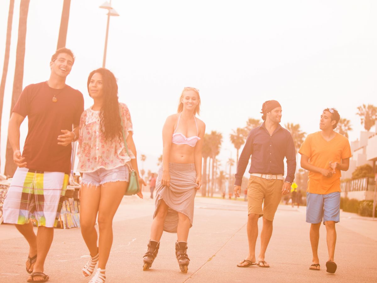 A group of five people are enjoying a sunny day, walking and rollerblading on a boardwalk lined with palm trees.