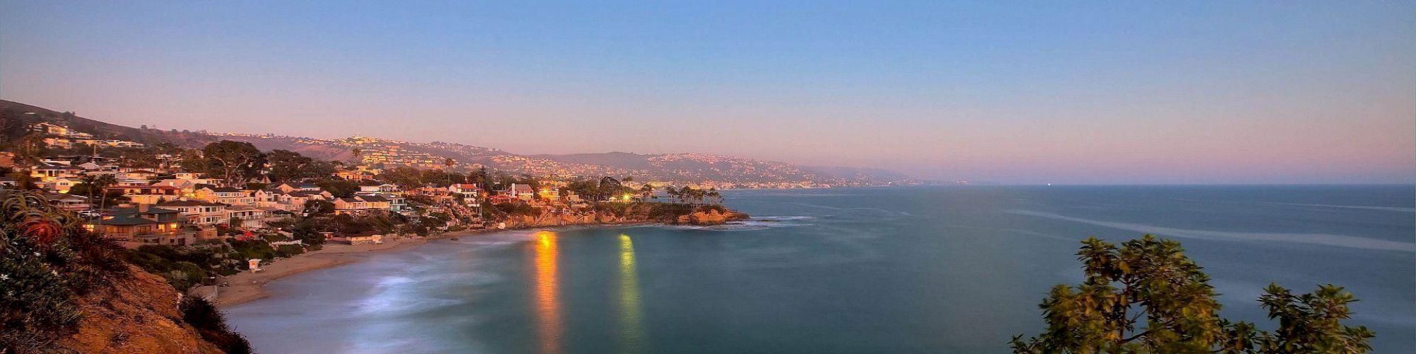 A coastal landscape at twilight showing a crescent moon, houses on a hill, calm sea, and reflections of lights on the water.