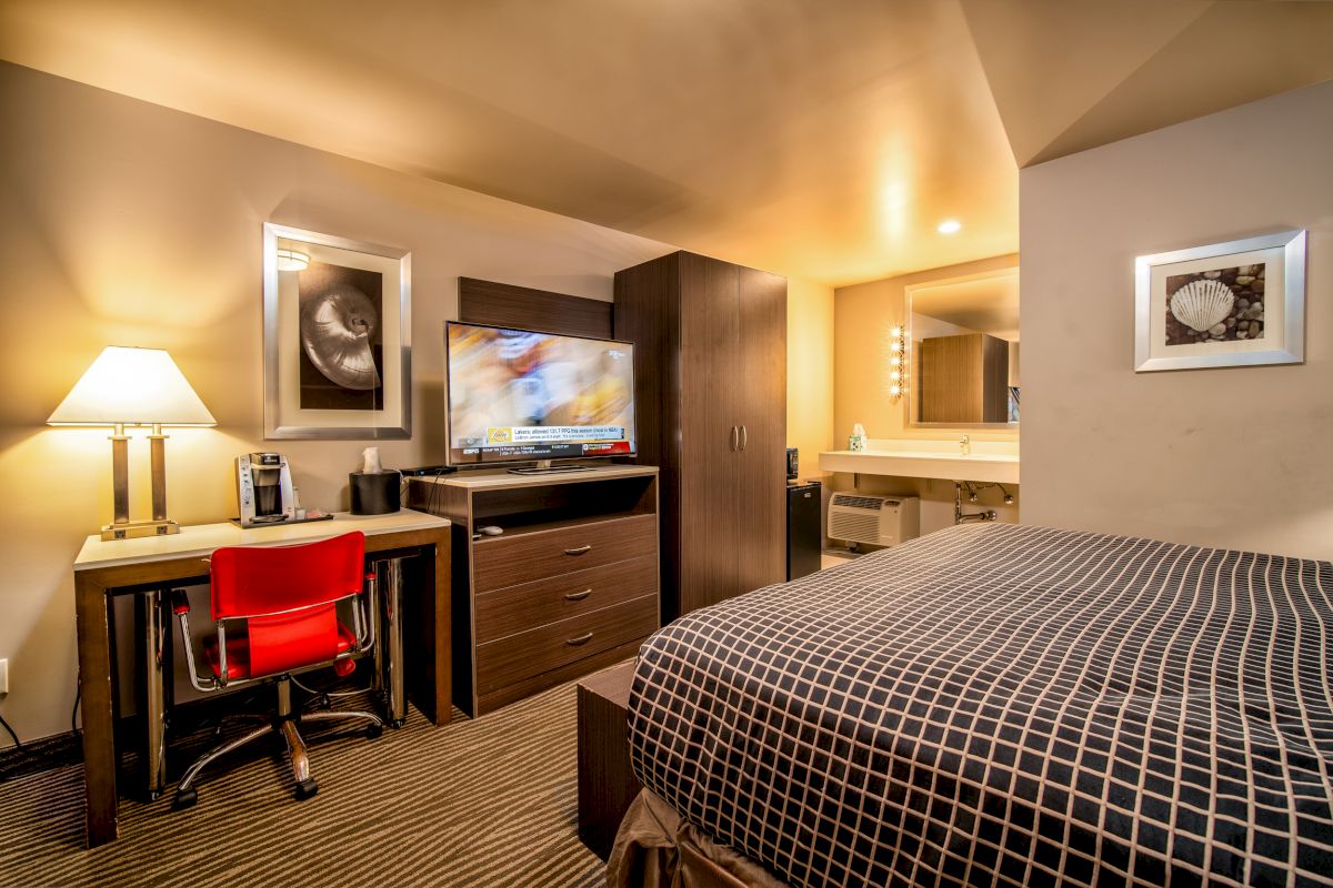 The image shows a hotel room with a bed, desk, lamp, red chair, TV, wardrobe, and a bathroom area featuring a mirror, sink, and lighted vanity.