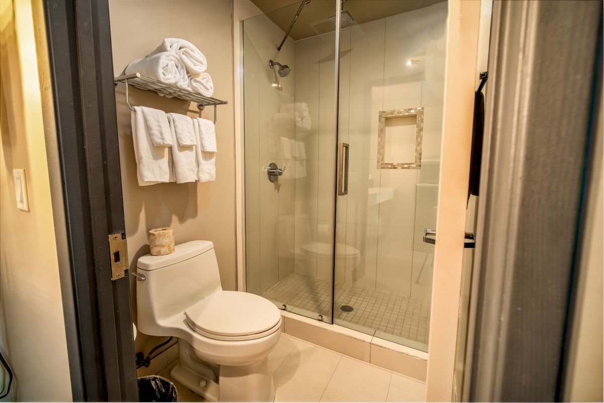 The image shows a modern bathroom with a toilet, a glass-enclosed shower, and a rack with folded towels and toiletries.