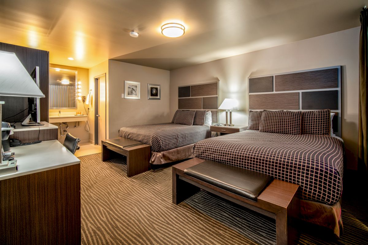 The image shows a hotel room with two double beds, a desk, a lamp, a bench at the foot of each bed, and an open bathroom area in the background.