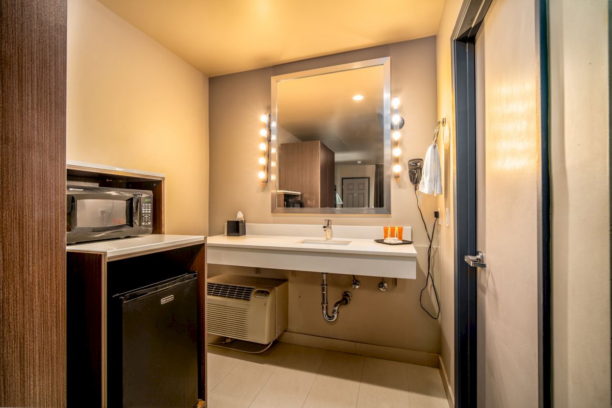 The image shows a hotel bathroom with a well-lit mirror, sink, hairdryer, and toiletries, alongside a microwave and a mini-fridge.