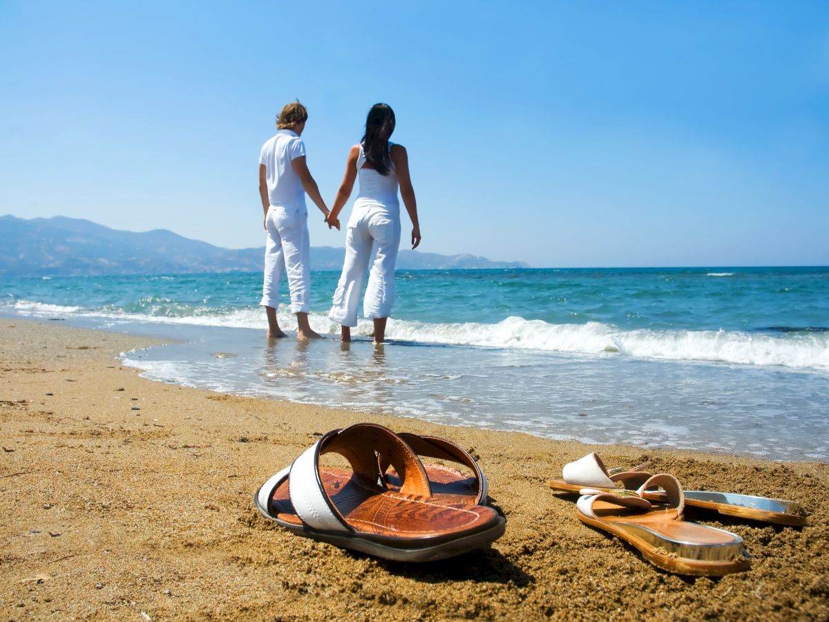 A couple holding hands walks along the shoreline, with sandals left in the sand in the foreground.