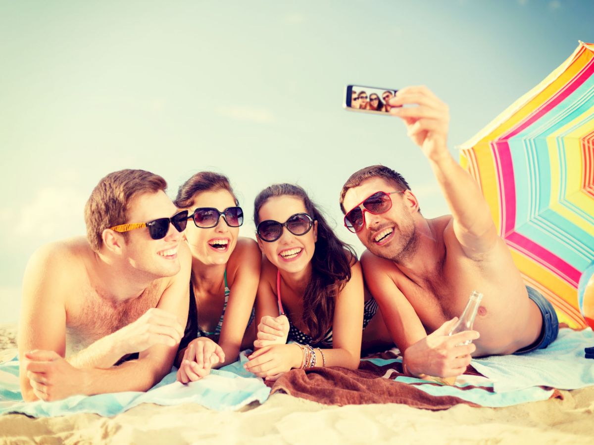 Four people lying on a beach, smiling, taking a selfie under a colorful umbrella. They all appear to be enjoying their time together.