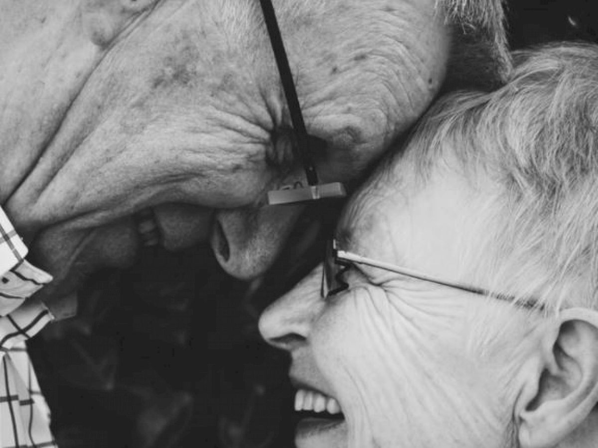 The image shows a close-up of an elderly couple touching foreheads, both smiling and appearing happy in a black-and-white photo.