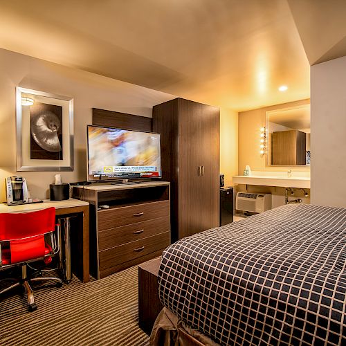 This image shows a hotel room with a bed, a desk with a lamp and chair, a television, and a bathroom area with a sink and mirror.