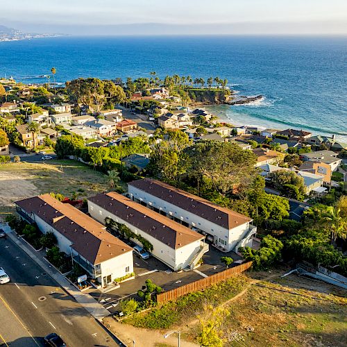 A coastal town featuring residential buildings, a nearby road, and a beautiful ocean view with a shoreline in the distance under a clear sky.