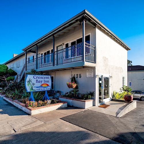 The image shows the Crescent Bay Inn, a two-story building with balconies, located on a sunny street with plants and a clear blue sky in the background.