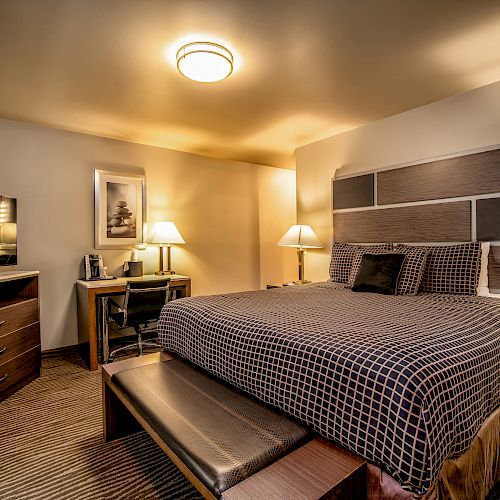 The image shows a cozy hotel room with a large bed, bedside lamps, a TV, a desk with a chair, and a bench at the foot of the bed.