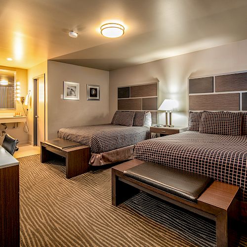 This image shows a hotel room with two double beds, a desk, a TV, a mirror, and a well-lit bathroom area in the background.