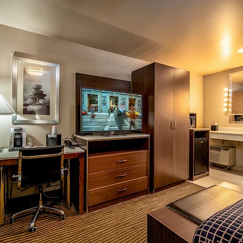 The image shows a modern hotel room with a desk, chair, lamp, TV, cabinets, and a sink area with a mirror illuminated by lights.