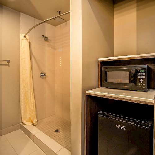 The image shows a bathroom with a shower area, a yellow shower curtain, towel on a rack, and a mini kitchen setup with a microwave and a mini-fridge.