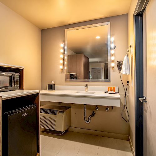 This image shows a hotel room bathroom with a vanity mirror, sink, towels, and a small microwave and refrigerator on the left.