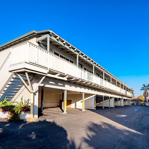 The image shows a multi-story apartment complex with exterior corridors and staircases, surrounded by a paved area and clear blue skies.