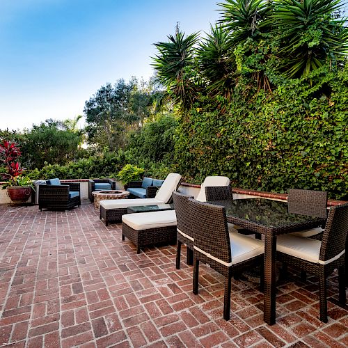 The image shows an outdoor patio with brick flooring, modern wicker furniture, and lush greenery. It includes lounge chairs and a dining table.