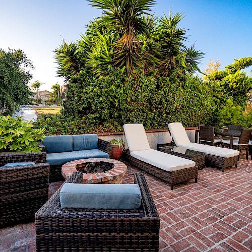 An outdoor patio area with wicker furniture, lounge chairs, a fire pit, and surrounded by lush greenery in a sunny setting.