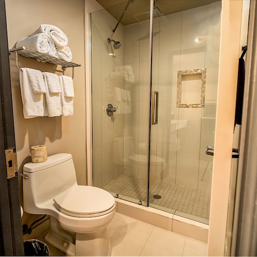 The image shows a bathroom with a toilet, towels on a rack, and a glass-enclosed shower with a modern design, featuring beige tile flooring.