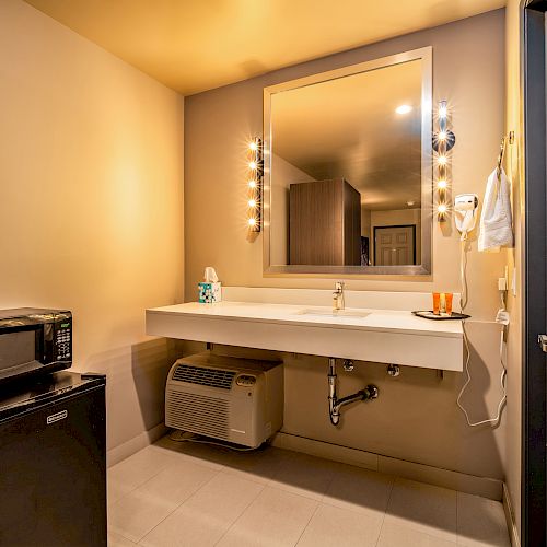 The image shows a hotel room vanity area with a large mirror, illuminated by lights, a countertop, a sink, a microwave, and a small refrigerator underneath.