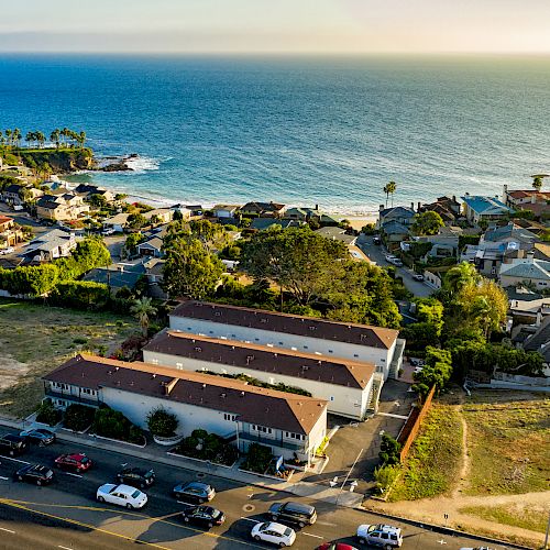 Aerial view of a coastal town with residential buildings, roads, and parked cars near a scenic ocean backdrop and sandy beach.