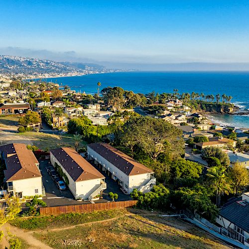 An aerial view of a coastal town features residential areas, a main road with cars, and the ocean with a distant shoreline under a clear sky.