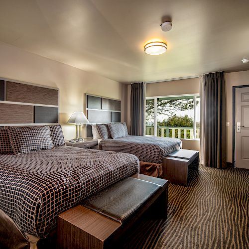 This image shows a hotel room with two double beds, a window with a view, bedside lamps, a dresser with a TV, and a door. The room is neatly arranged.