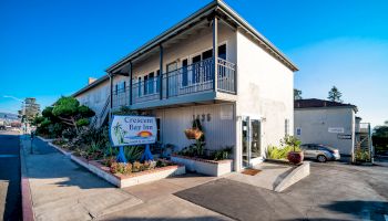 The image shows a two-story building with a sign reading "Crescent Bay Inn" surrounded by plants and a parking area on a sunny day.