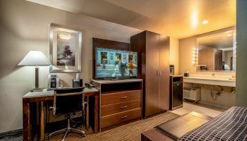 A neatly organized hotel room with a desk, a chair, a lamp, a flat-screen TV, a wardrobe, a mirror, and a sink area, creating a cozy atmosphere.