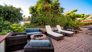 This image shows an outdoor patio with wicker furniture, including lounge chairs, a fire pit, and a dining table, surrounded by lush greenery.