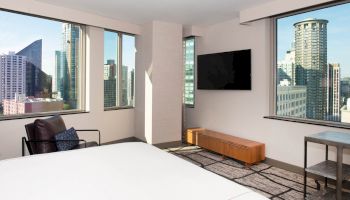 A modern hotel room with large windows showing a cityscape view, a bed, a chair, a wall-mounted TV, and a small table with a lamp.