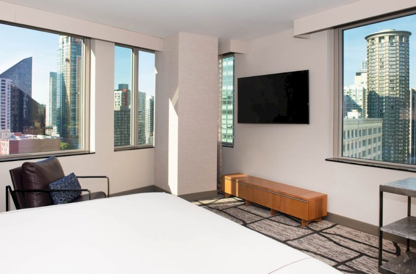 A modern hotel room with large windows showing a cityscape view, a bed, a chair, a wall-mounted TV, and a small table with a lamp.
