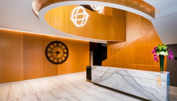 A modern lobby with a marble reception desk, wooden spiral staircase, large black clock on the wall, and flower arrangement on the counter.