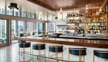 The image shows a modern, stylish bar with blue and gold stools, a well-stocked bar, and large windows offering a city view, under a wooden ceiling.