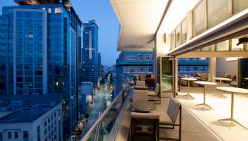 A modern cityscape at dusk, highlighting a stylish terrace with seating and tables, overlooking a street lined with tall buildings.