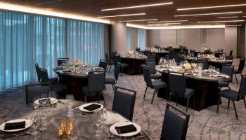 The image shows a modern, elegantly set dining room with round tables, black chairs, neatly arranged place settings, and dim lighting.