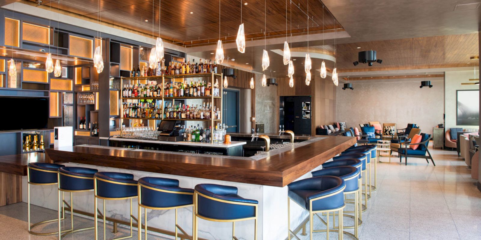 A modern bar with a well-stocked shelf, stylish hanging lights, and blue-and-gold bar stools. The background has lounge seating with a large window.