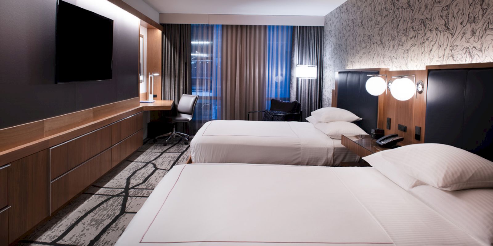 The image shows a modern hotel room with two double beds, a desk, TV, and stylish decor. The room is well-lit with lamps and natural light.