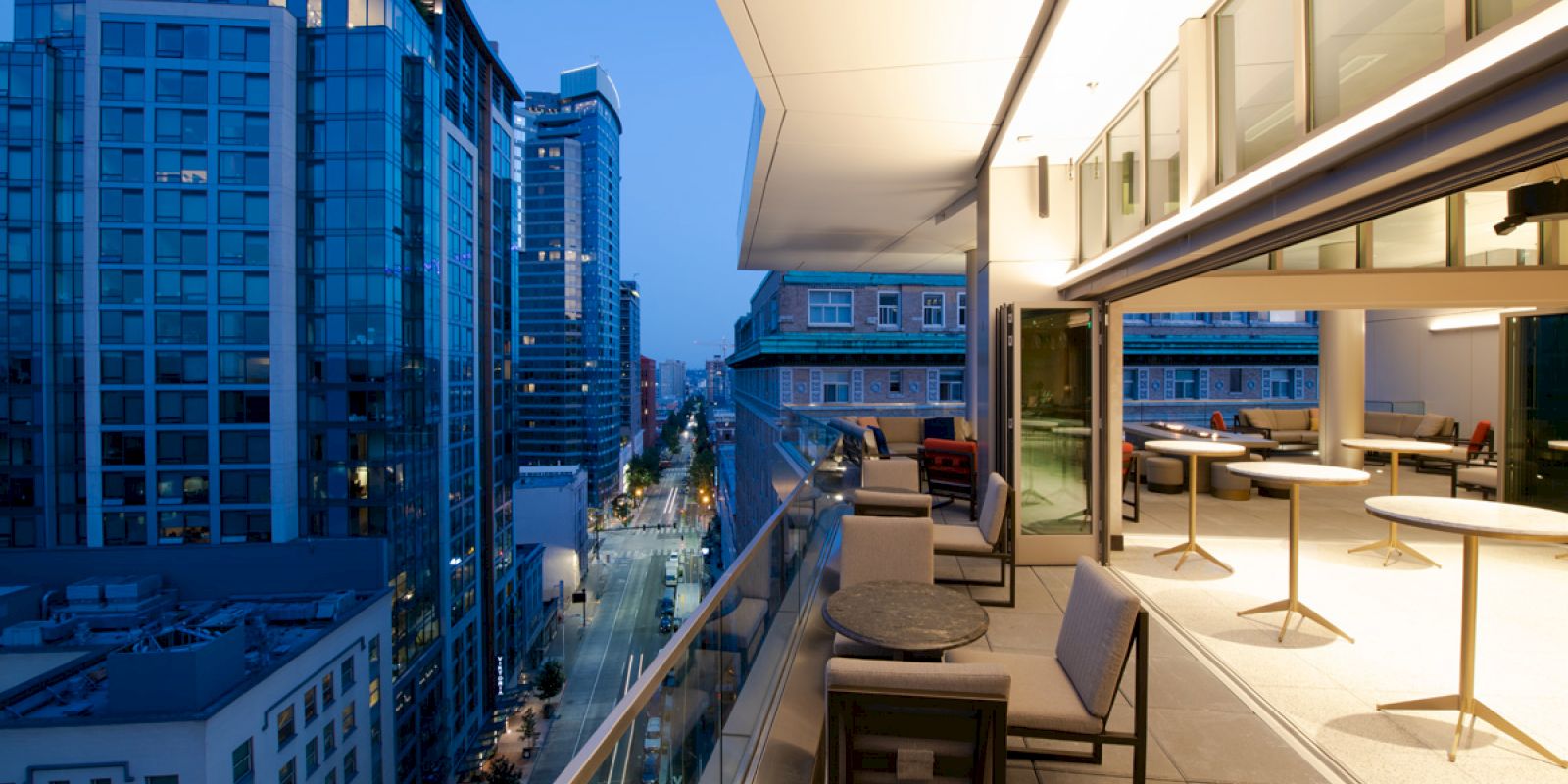 A modern urban balcony at dusk with tables, chairs, and cityscape views of tall buildings and streets below, illuminated by city lights.