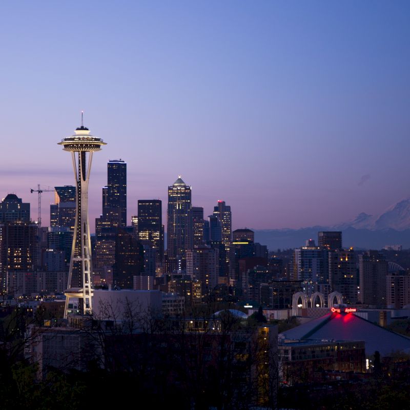 It’s a cityscape of Seattle at dusk, featuring the Space Needle and various buildings, with Mount Rainier visible in the background.