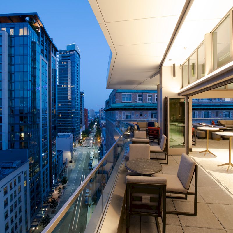A modern balcony overlooks a city street with tall buildings, featuring outdoor seating and adjacent glass-paneled indoor spaces, illuminated at dusk.