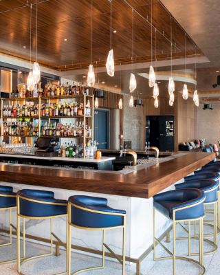 The image shows a modern bar with blue stools, pendant lights, and a well-stocked liquor shelf behind the counter, in a spacious, stylish setting.