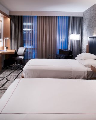 A modern hotel room with two neatly made beds, a large TV, a desk with a chair, floor lamps, and full-length curtains.