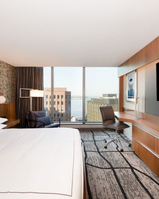 A modern hotel room features a large bed, a desk with a chair, a flat-screen TV, and a window with a city view.